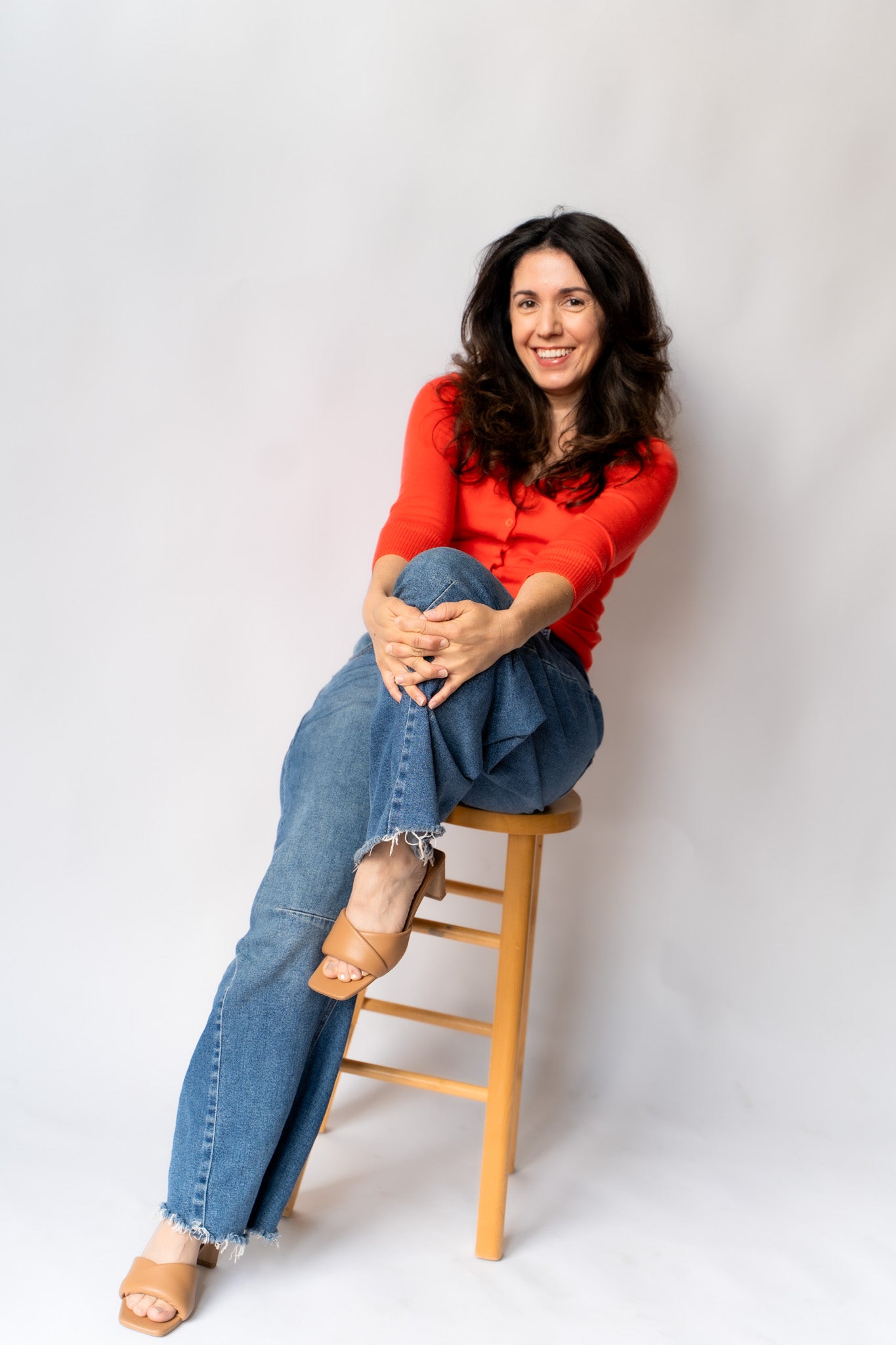 Woman sitting on a wooden stool wearing a red shirt and blue jeans against a white background