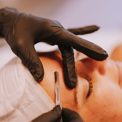 Person receiving a facial treatment with gloved hands using a dermaplaning tool.